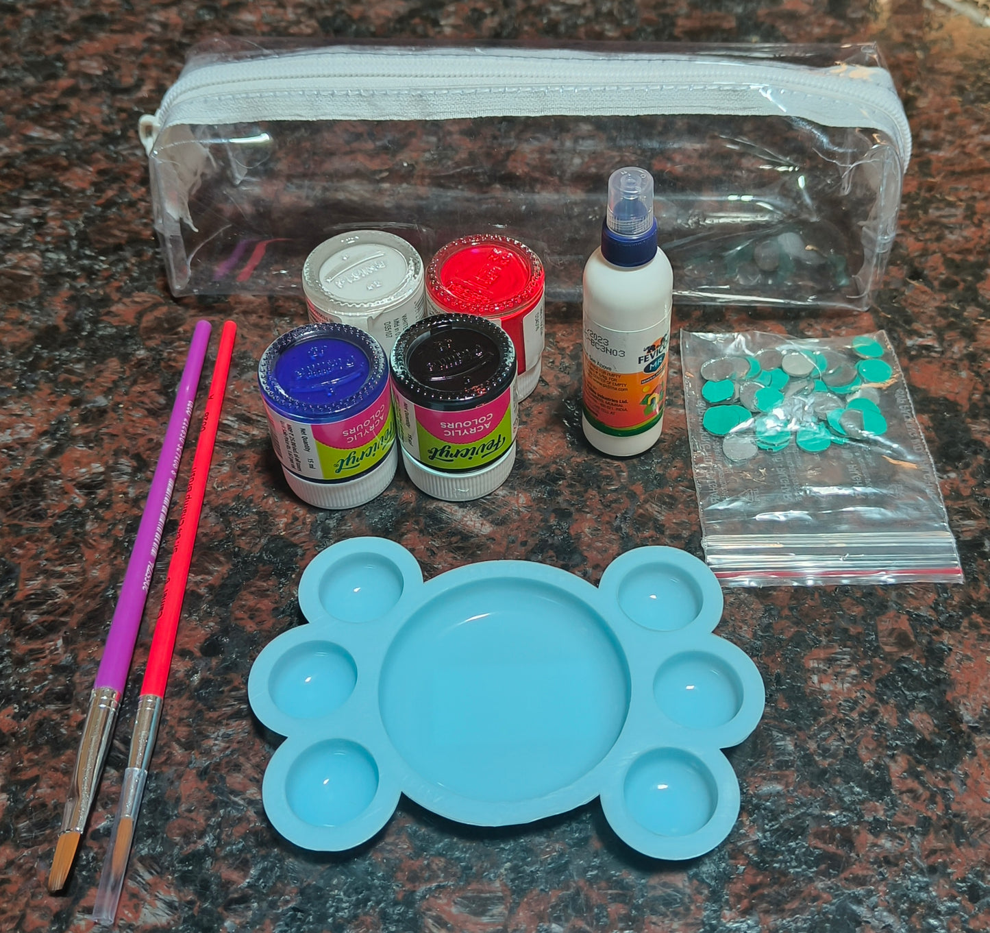 4 acrylic paint bottles (red, white, blue, and black), a blue palate, one purple flat brush, one red round brush, a fevicol bottle, a pouch with round mirrors, and an empty transparent pouch - all placed on a granite background.