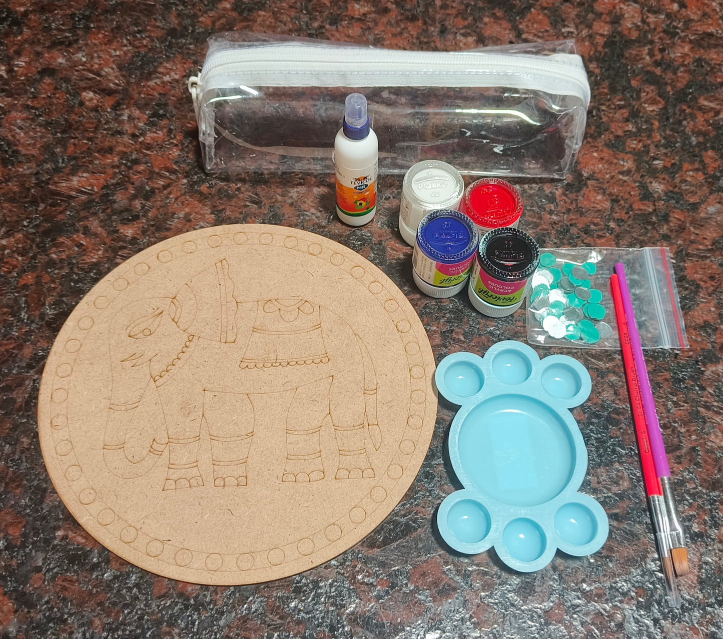 A round MDF board with elephant design, 4 acrylic paint bottles (red, white, blue, and black), a blue palate, one purple flat brush, one red round brush, a fevicol bottle, a pouch with round mirrors, and an empty transparent pouch - all placed on a granite background.
